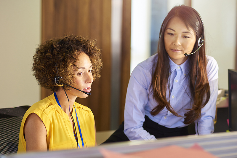 Health management services - a young call centre representative greets a caller in a large open plan office as her supervisor watches over her shoulder and helps her through the call.