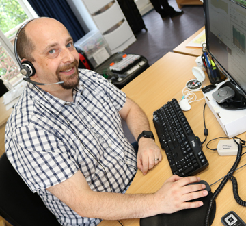 A man sitting at a computer wearing a headset and smiling at the camera