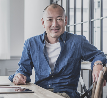 Man sitting at a table with his arm resting on the back of the chair and turned towards the camera