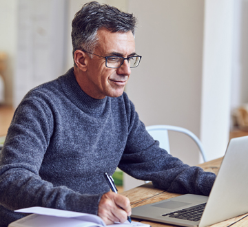 Man sitting at a table looking at a laptop and holding a pen over paper