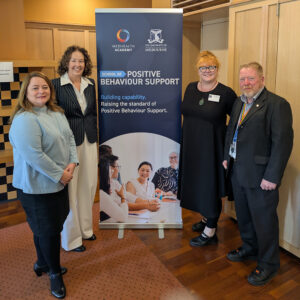 A photo of four people in front of a MedHealth Academy and the University of Melbourne banner, all smiling at the camera. They are Cara Newman, Director Clinical Services, Outcomes Connect Australia; Amanda Wood, General Manager, Real Therapy Solutions, Leonie Harding, Learning & Development Lead, Outcomes Connect Australia, and Professor Keith McVilly