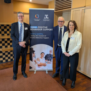 Three people are standing in front of a banner which says School of Positive Behaviour Support on it. They are smiling. Their names are Tim Morphy, CEO MedHealth; Peter Steadman, Chief Medical Officer, MedHealth; Dorothy Frost, Head of Clinical Capability and Innovation, MedHealth.